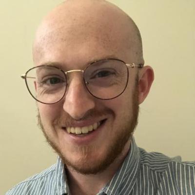 image of smiling white male with glasses and a reddish beard. Raymond is wearing a blue and white striped collared shirt 